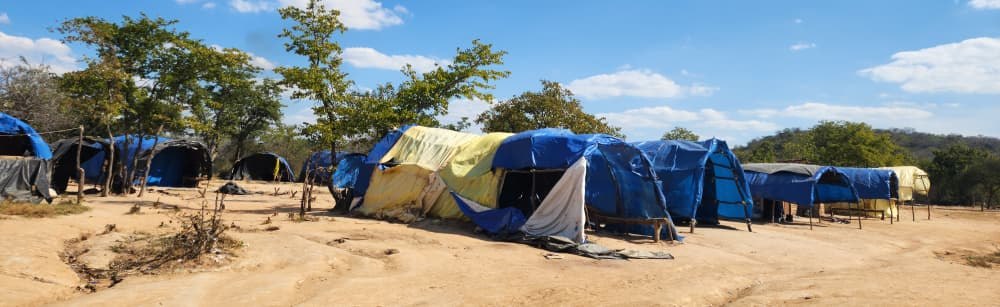 Rows of tarpaulin and pole shelters at an artisanal mining settlement on open ground in Matabeleland South, Zimbabwe, with trees and hills behind