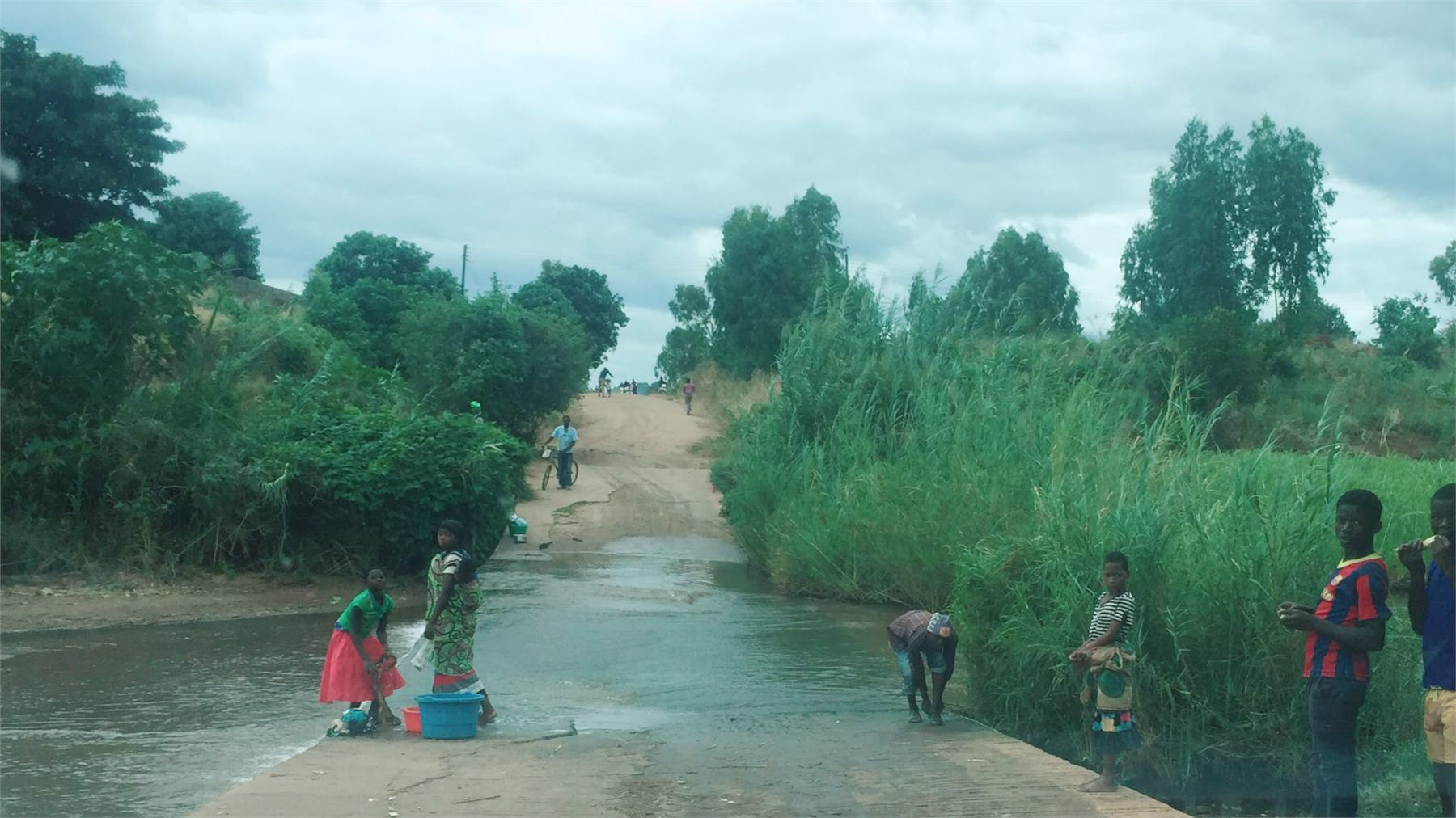 Community members crossing a flooded causeway on a rural route, with a cyclist and pedestrians fording the water