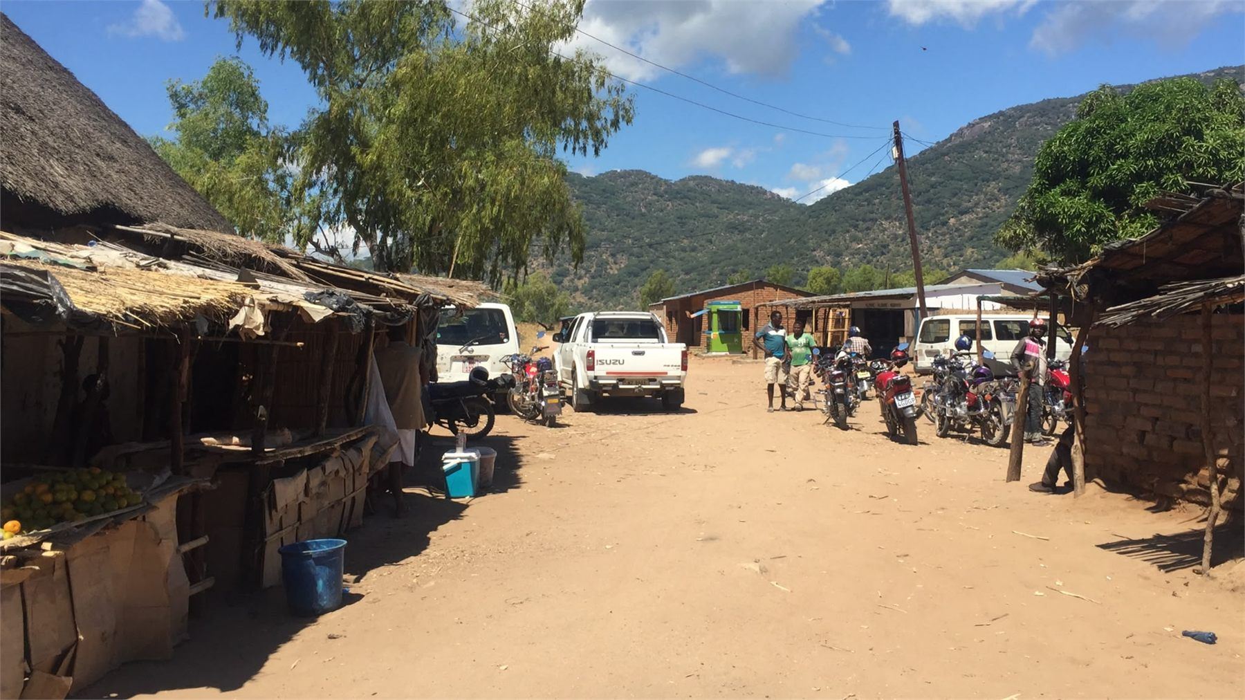 Rural trading centre in Malawi with vehicles, motorcycles and market stalls against a mountain backdrop