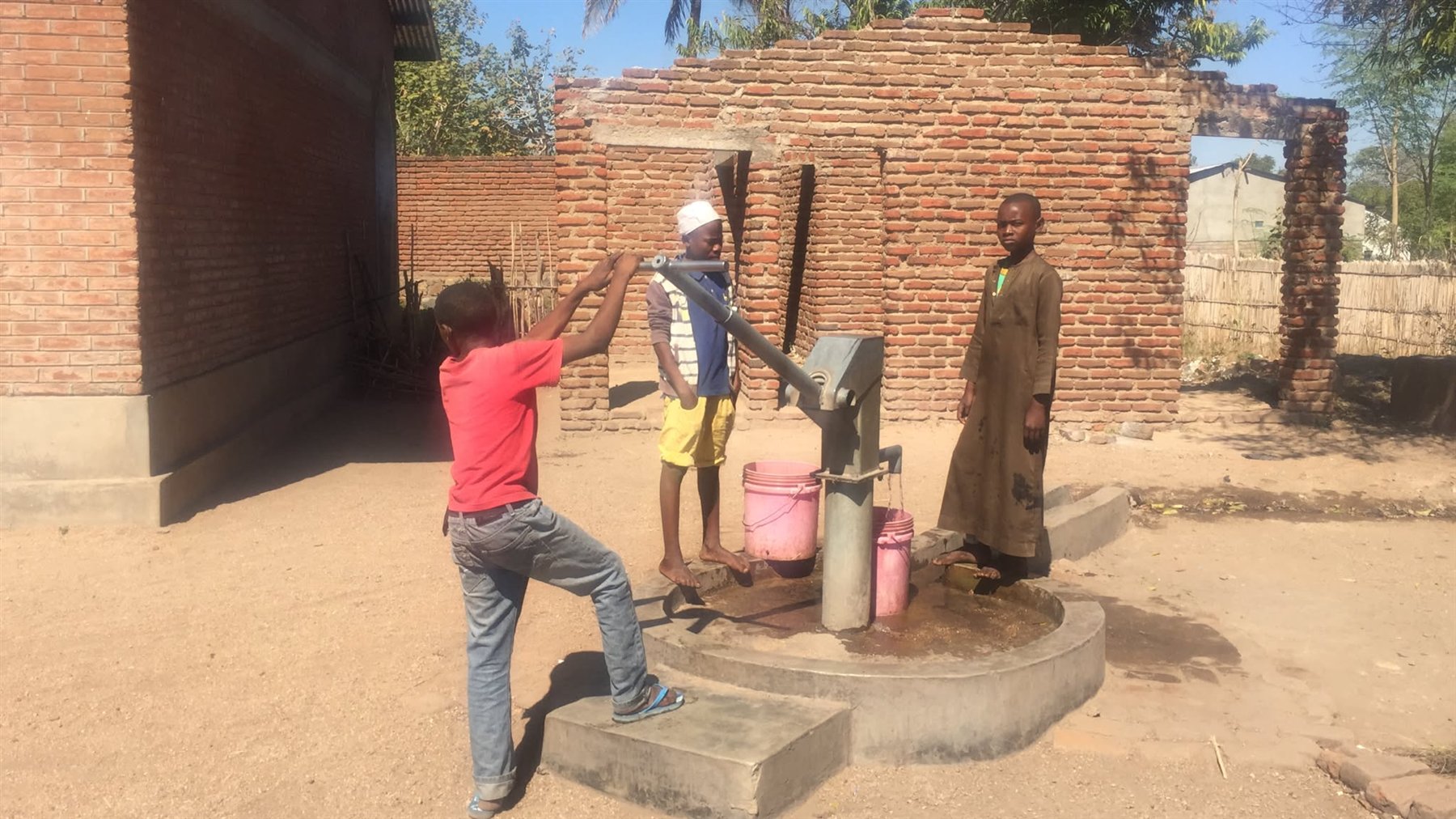 Children filling buckets at a community handpump beside a brick building in a village in Malawi