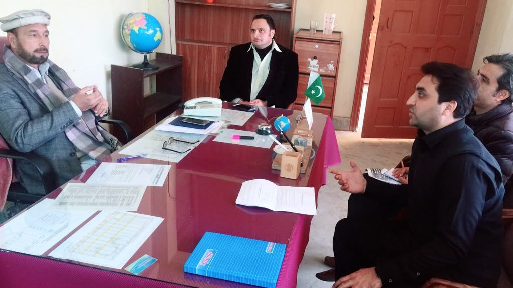 Four people seated around a desk spread with printed service registers during an interview at a district office, with a Pakistan flag on the desk