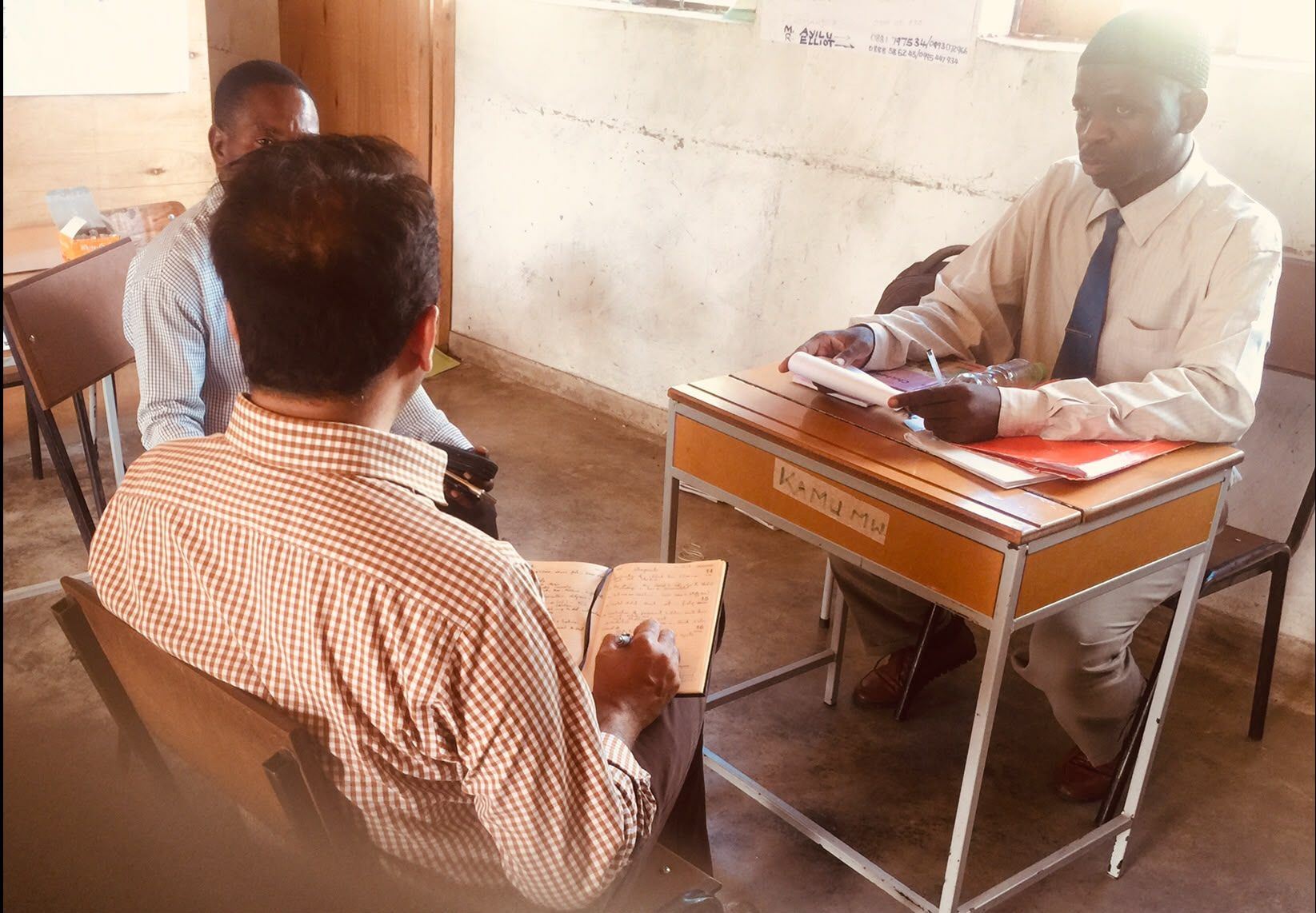 PMEye specialist taking handwritten notes during a key informant interview with a head teacher in a school classroom in Malawi