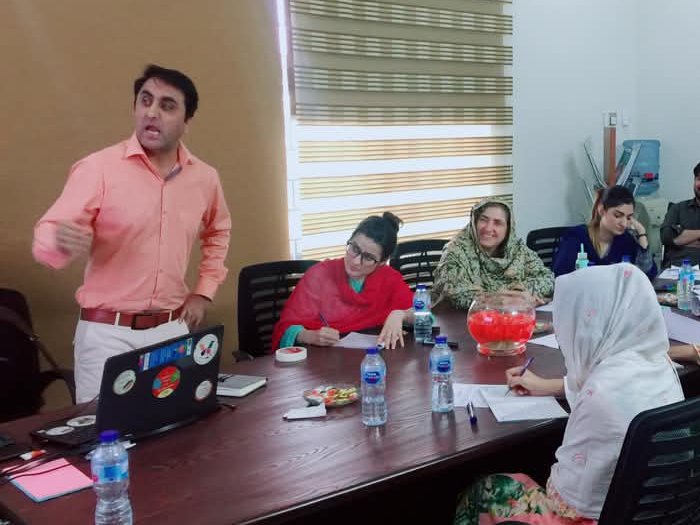 PMEye trainer standing and presenting to programme staff seated around a conference table, taking notes, during a project management training in Peshawar
