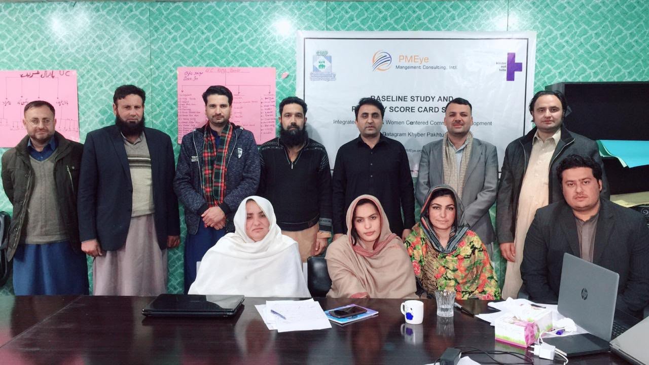 Enumerator training group with PMEye and partner banners on the wall, men standing and women seated at a conference table, Batagram
