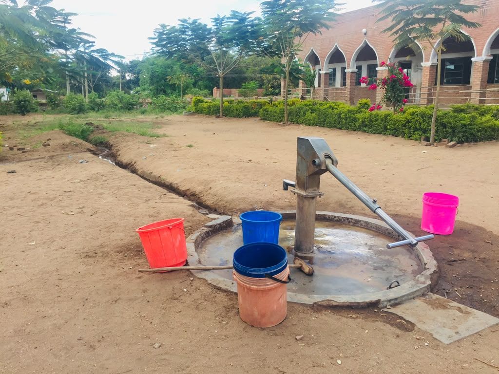 Hand pump on a concrete apron with drainage channel and collection buckets, beside an institutional building