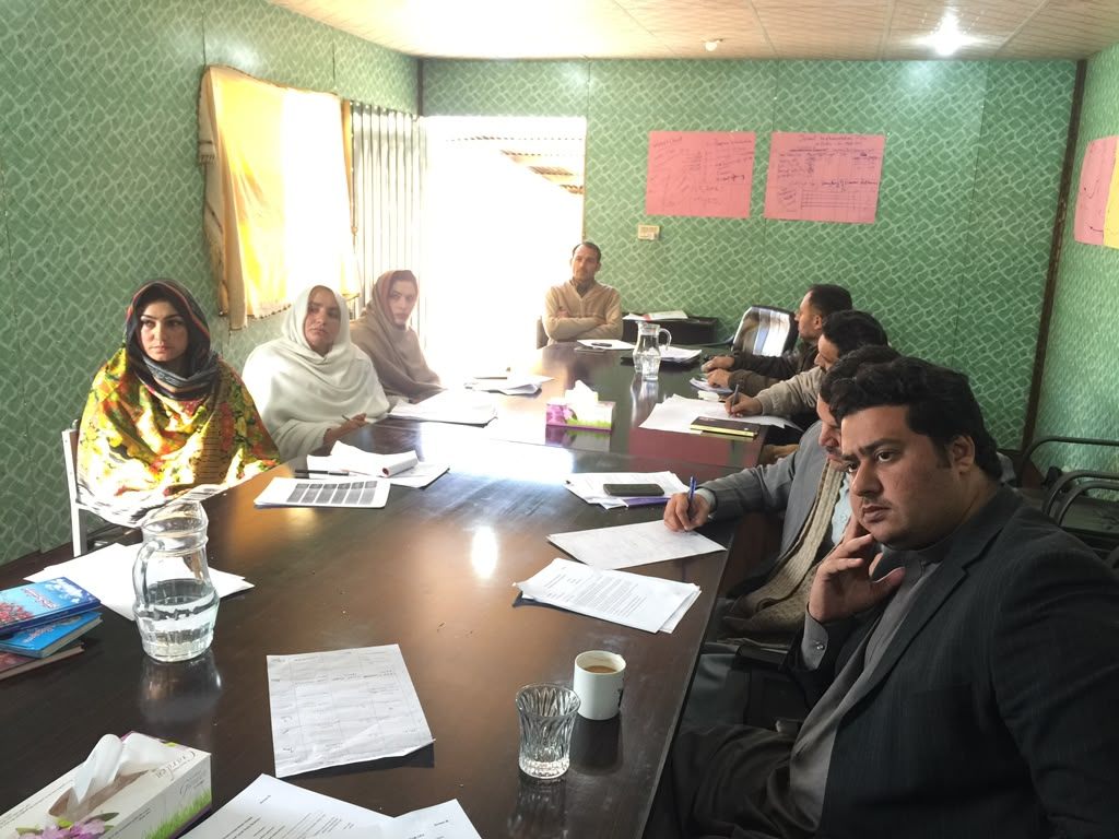 Stakeholder working session with participants seated around a table reviewing printed documents, flipcharts on the wall